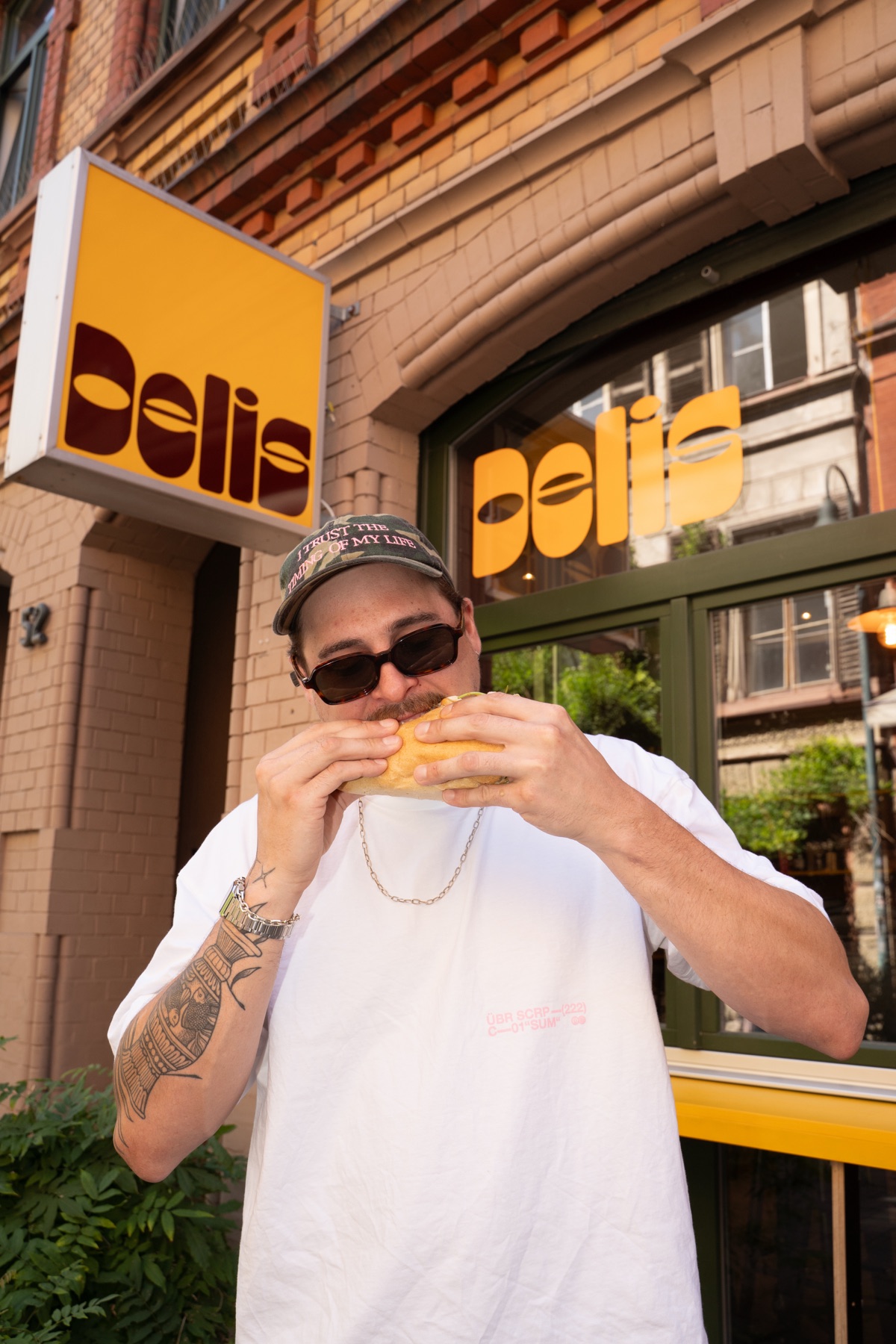 A guest taking a big bite of a sandwich in front of the yellow DELIS sign