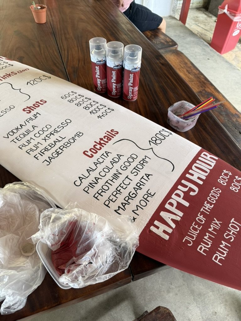 Painting the happy hour menu onto a surfboard, red paint and brushes on a table